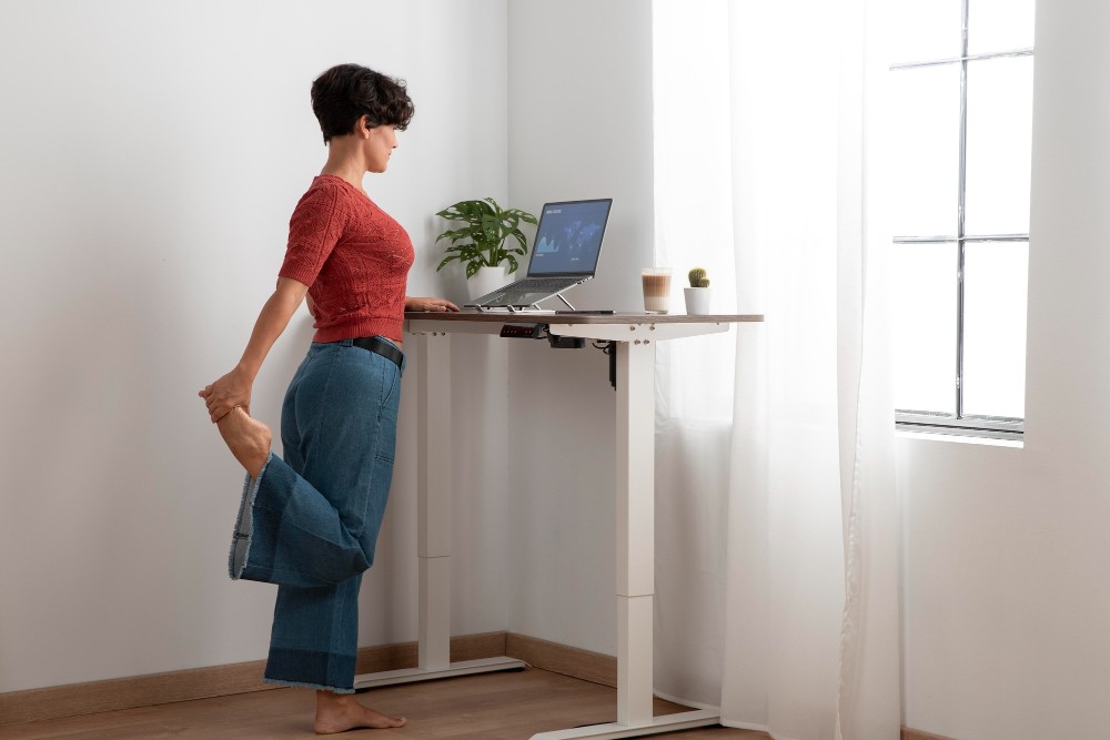 Abbildung: Eine Person steht an einem höhenverstellbaren Schreibtisch. Ein Bein winkelt sie nach hinten ab und umfasst ihren Fuß.