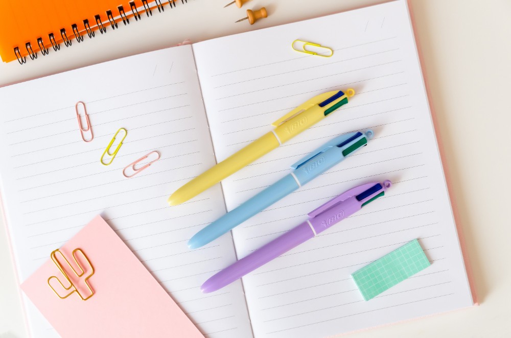 Drei verschiedenfarbige Kugelschreiber liegen auf einem Schreibblock: hellgelb, hellblau und helllila.
